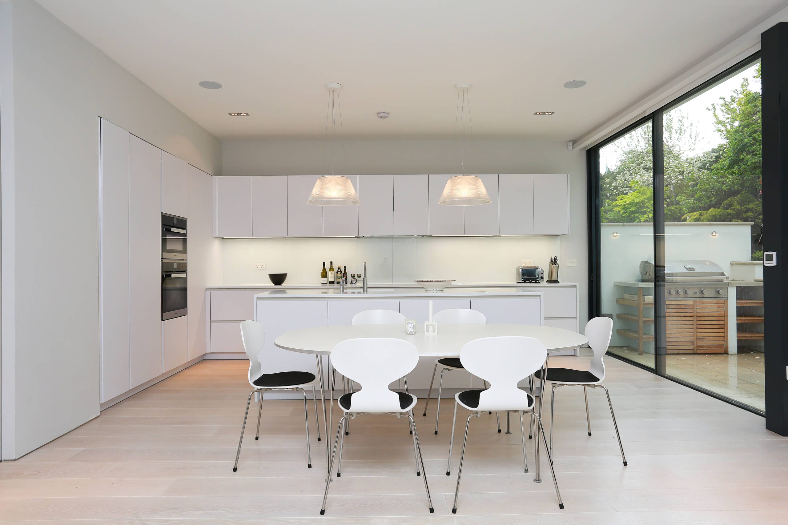 white-matt-kitchen-fitted-in-Liverpool Modern white matt handleless kitchen fitted in Liverpool, featuring a central island, integrated appliances, minimalist dining area, and garden views through large sliding doors.